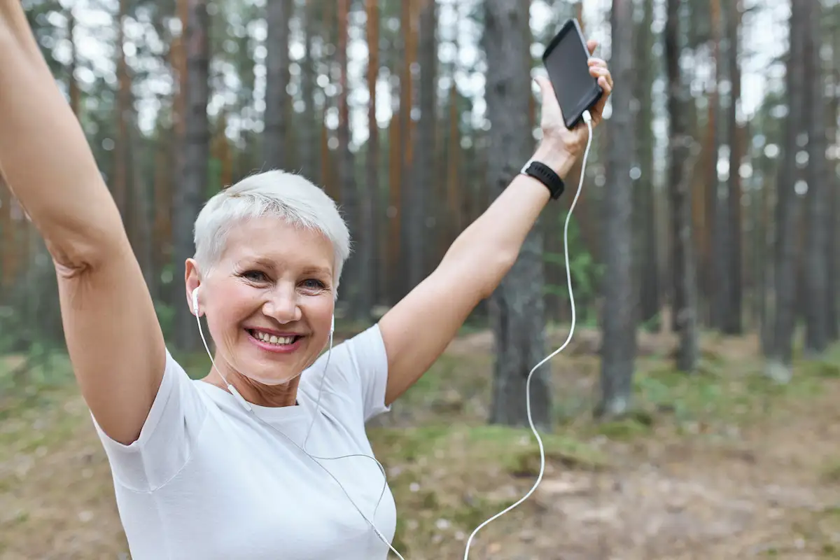 INÍCIO Energetic Cheerful Retired Female With Fit Slim Body Posing Outdoors Earphones Raising Hands Holding Cell Phone